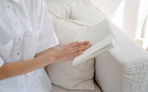 High of crop unrecognizable female wearing white shirt sitting on sofa and reading notepad