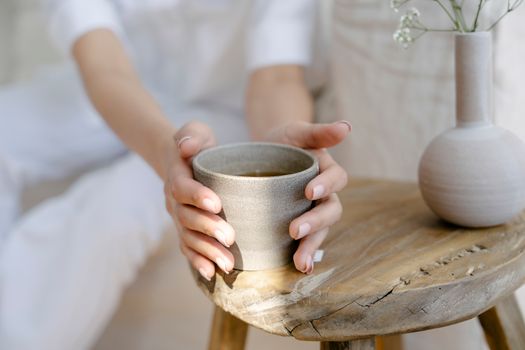 Crop anonymous female with ceramic cup of herbal tea placed on round table near vase of twigs at home