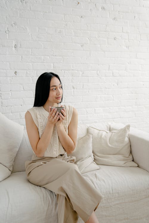 Serene ethnic female sitting on couch with cushions and enjoying hot drink at home against brick wall