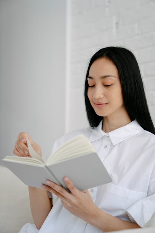 Free Dark haired ethnic female with long hair reading interesting novel while sitting on sofa against brick wall Stock Photo
