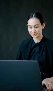 Smiling woman in black attire working on a laptop, embodying focus and positivity in an indoor office setting.