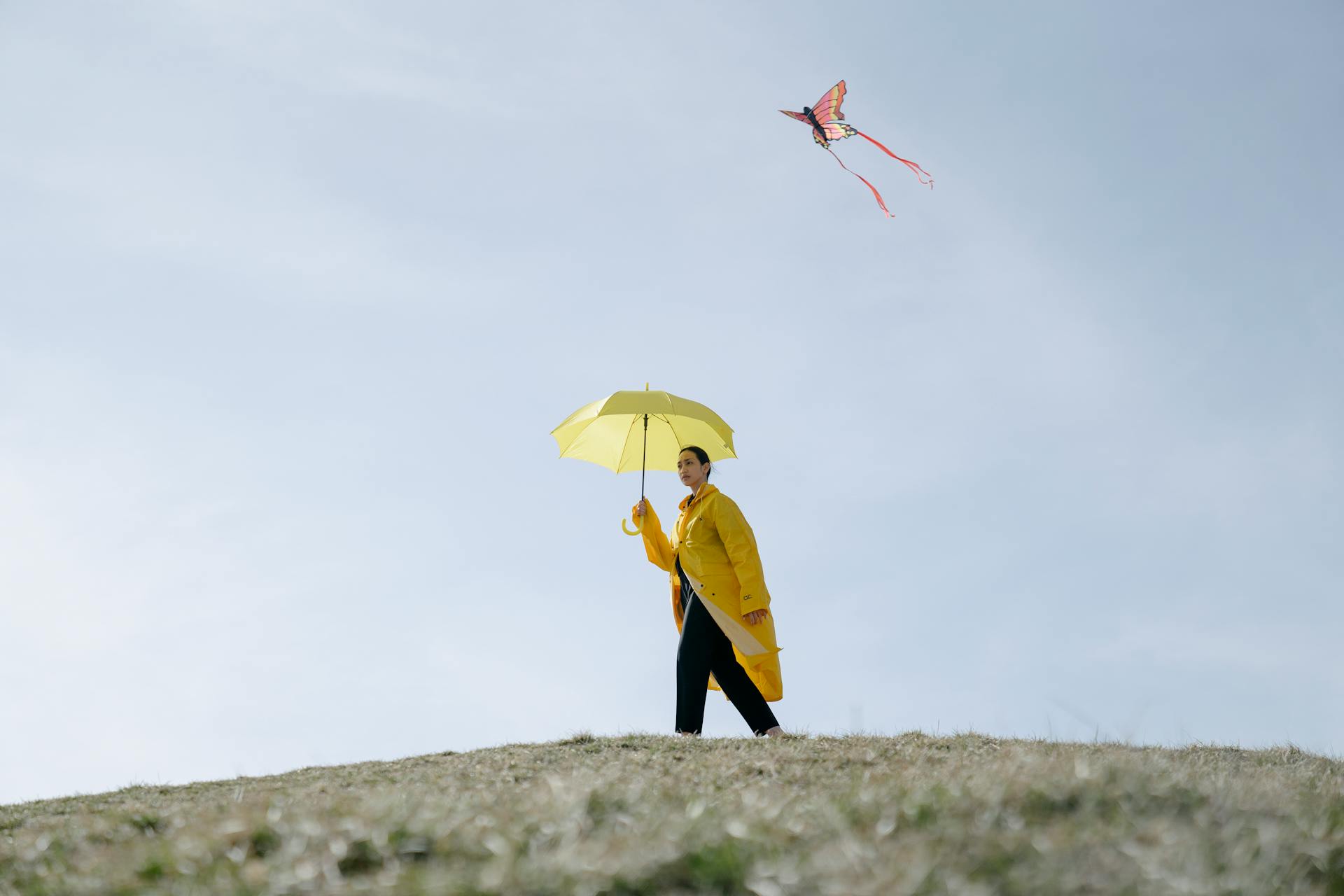 A woman in a yellow jacket walks on a hill with a yellow umbrella and a kite flying overhead.