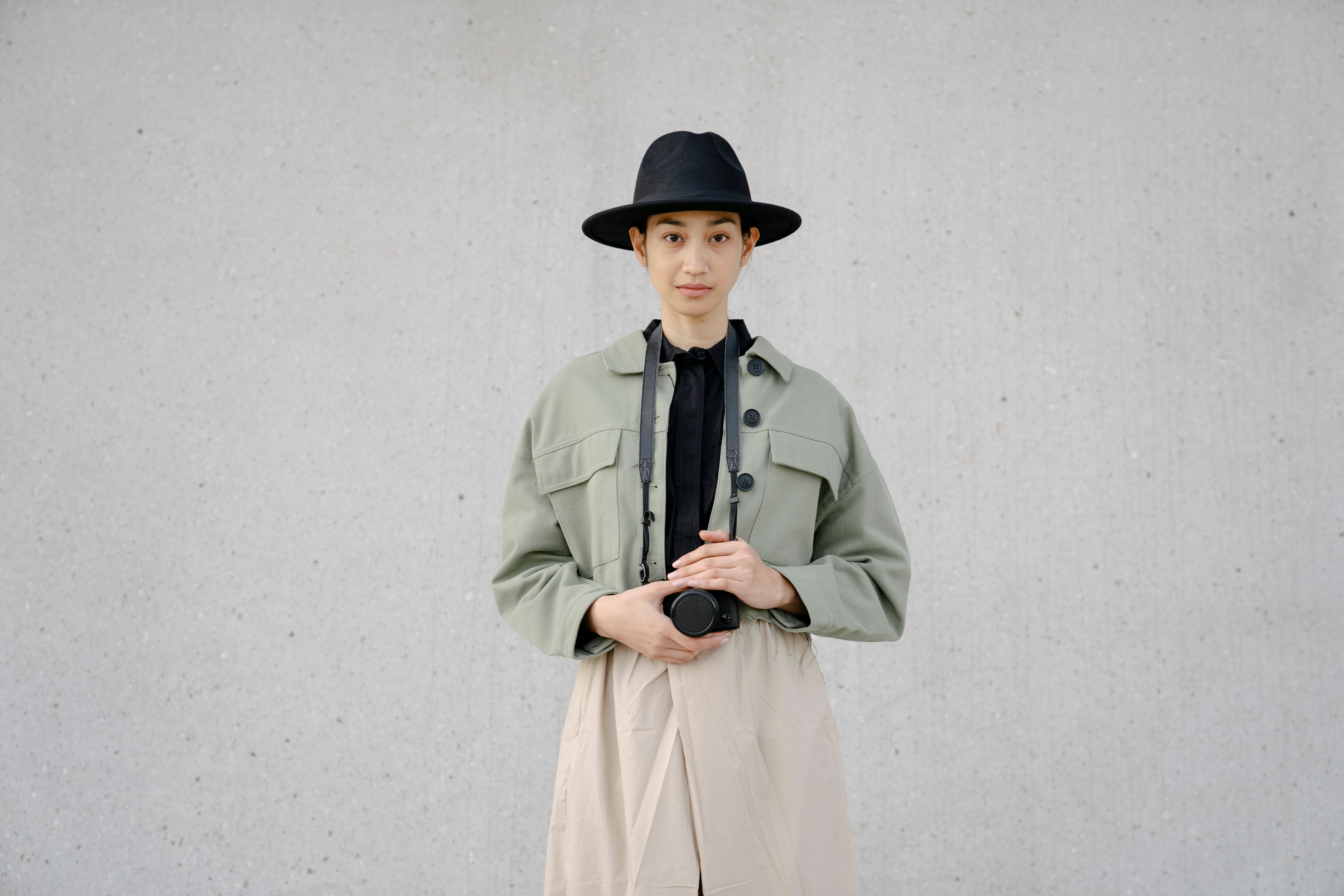 Fashionable woman with a camera, wearing a stylish hat and coat, standing against a minimalist background.