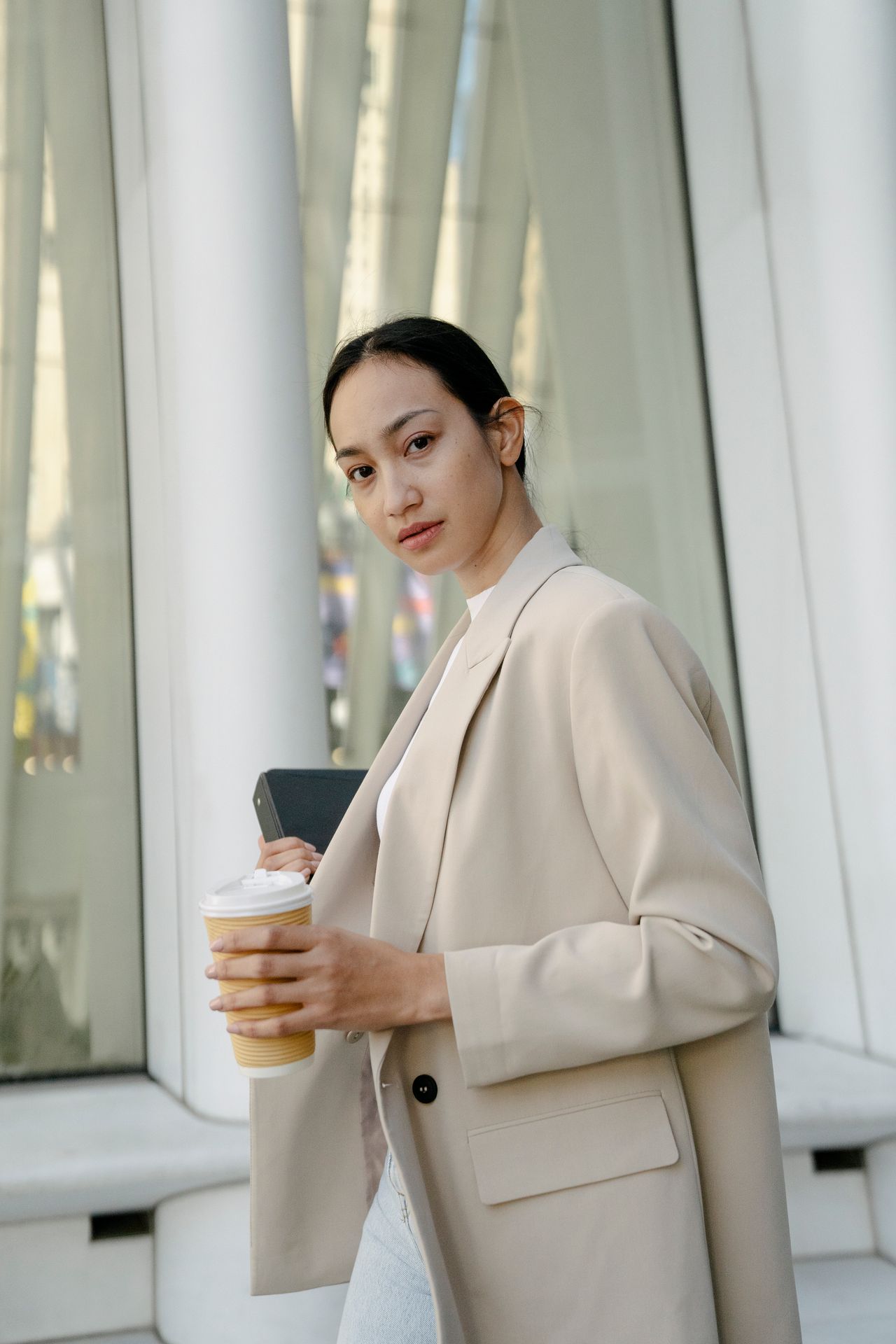 Woman in a beige blazer holding a coffee cup and a tablet while standing outdoors.