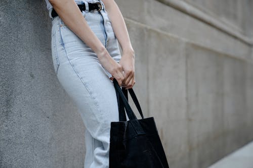 Crop anonymous female in casual jeans and with black eco friendly bag standing near wall in city street