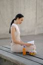 Side view of young ethnic female reading textbook on bench with bottle of drink and fruit salad in town