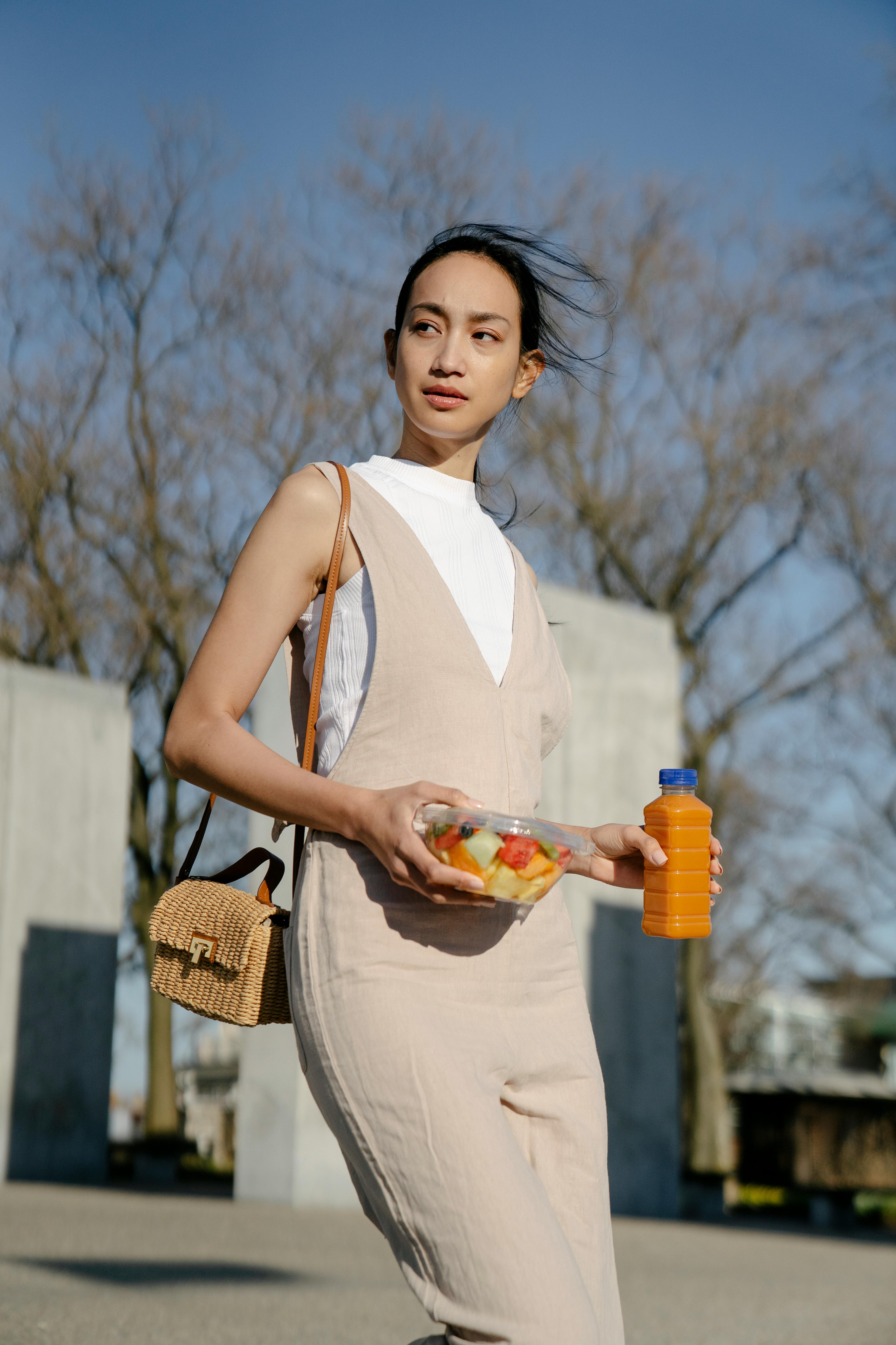 Young Asian female with disposable container of fruit salad and bottle of drink looking away while strolling in city