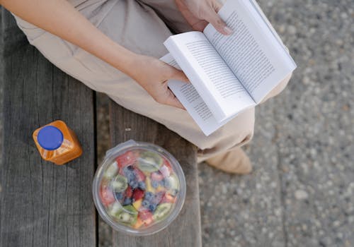 From above of crop unrecognizable female reading textbook on street bench with refreshing drink and fruit salad in takeaway container