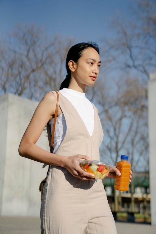 Trendy young Asian female with disposable box of fruit salad and bottle of beverage looking forward while walking in city