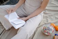 High angle of crop anonymous female reading textbook on crumpled fabric with disposable box of fruit salad and fresh tangerines