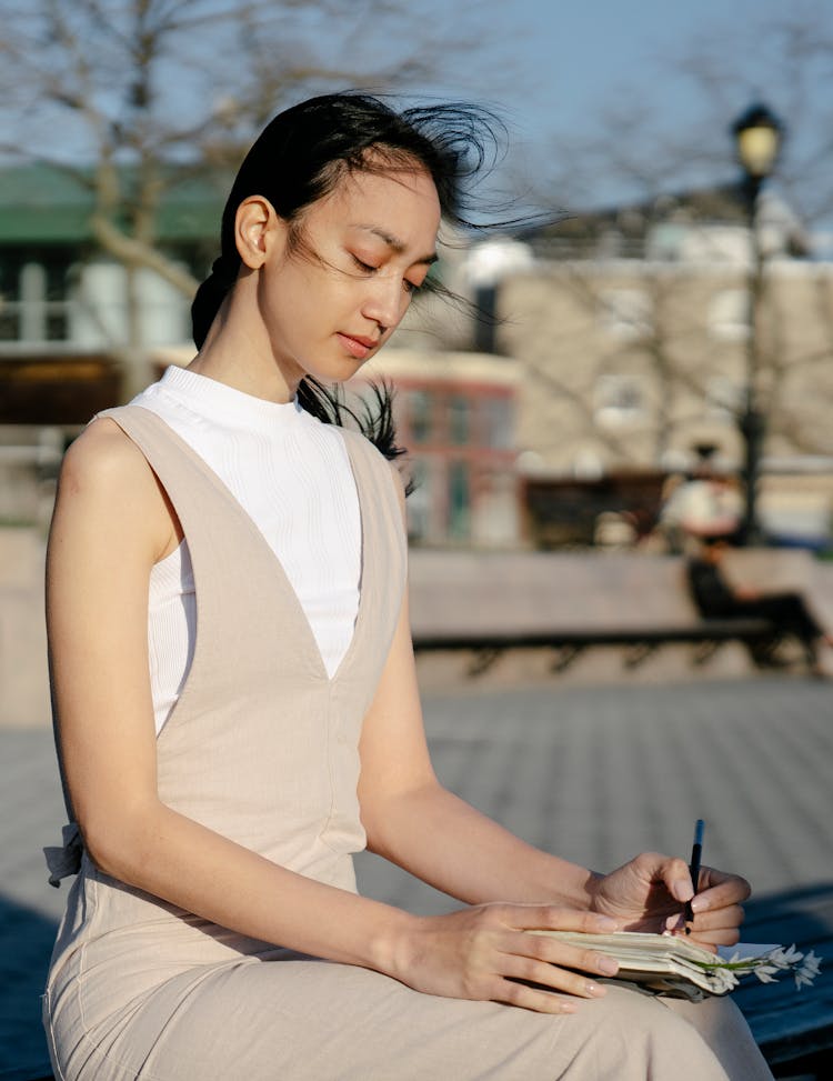 Asian Woman Writing In Diary On Urban Bench