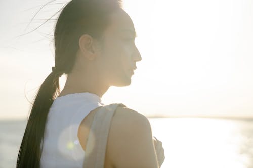 Back view of reflective young Asian female admiring glowing ocean while looking away in sunlight