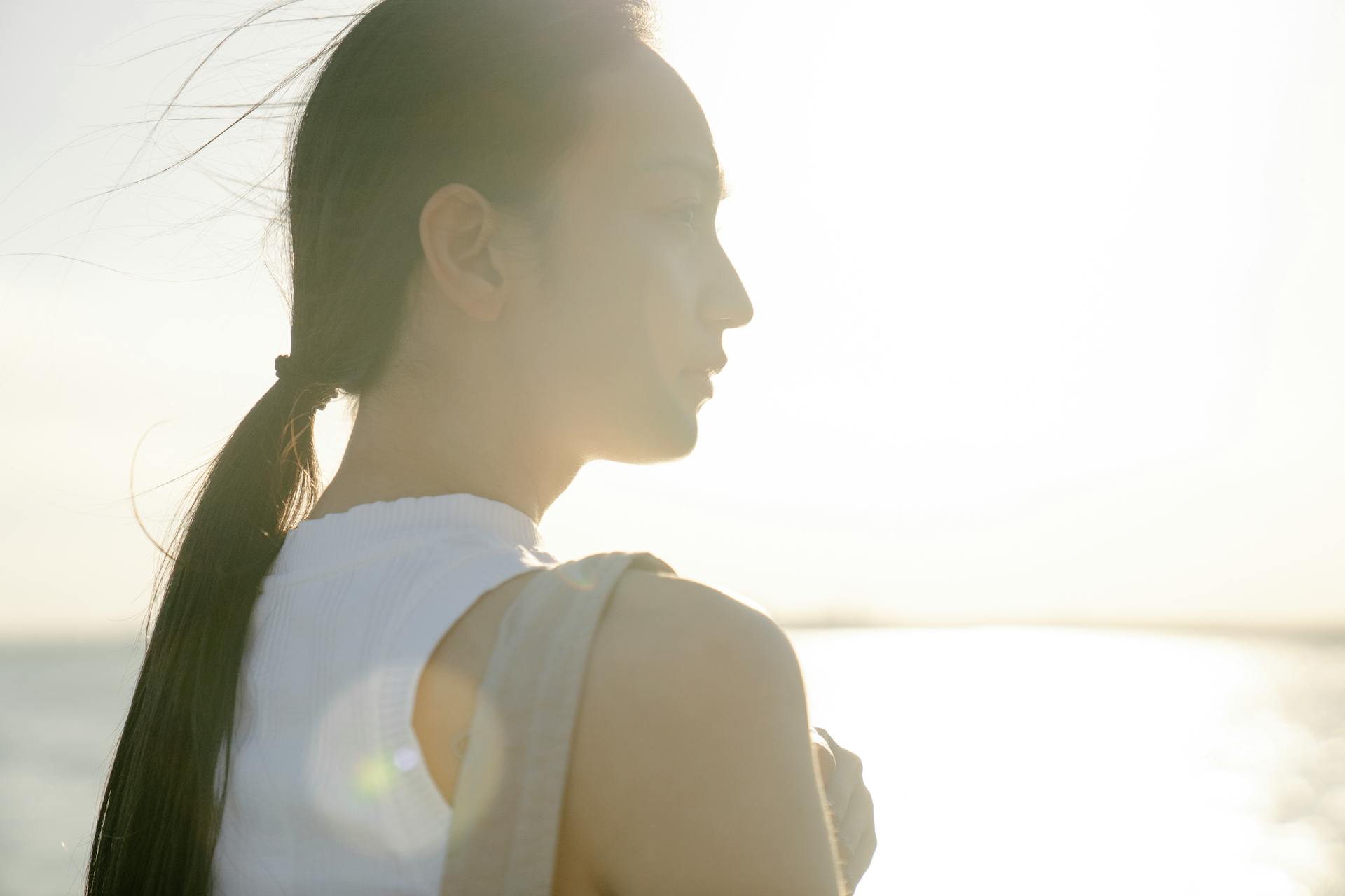 Back view of reflective young Asian female admiring glowing ocean while looking away in sunlight