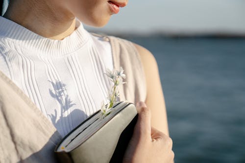 Crop unrecognizable gentle female with blossoming flower in diary against river on sunny day
