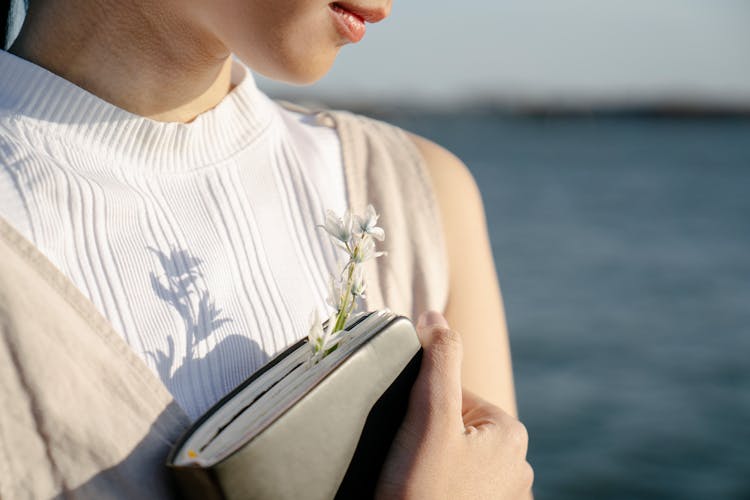 Crop Romantic Woman With Blooming Flower In Notebook Against Lake