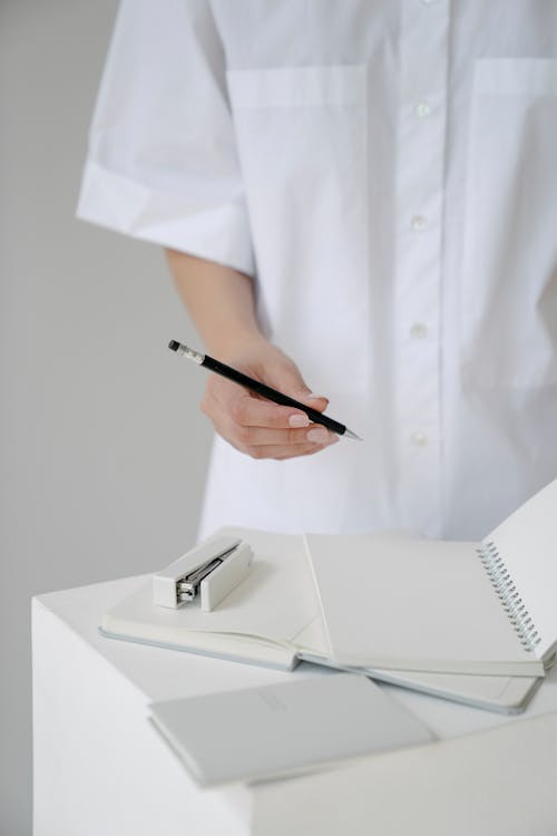 Free Crop unrecognizable female medic in white uniform with pencil and empty agendas on stand in hospital on gray background Stock Photo