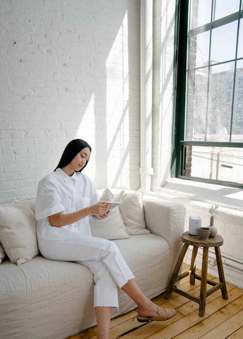 Young Asian lady with long dark hair in casual outfit sitting on comfortable sofa near window and reading interesting book during weekend at home