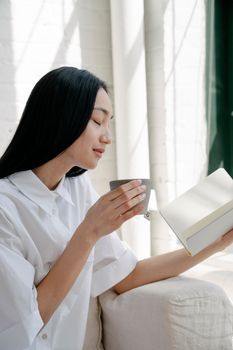 Content young ethnic female with long hair in casual clothes sitting on sofa and enjoying cup of aromatic coffee with closed eyes while reading book in sunny morning at home