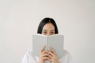 Young ethnic female student with long dark hair covering face with notebook while preparing for exams against white background