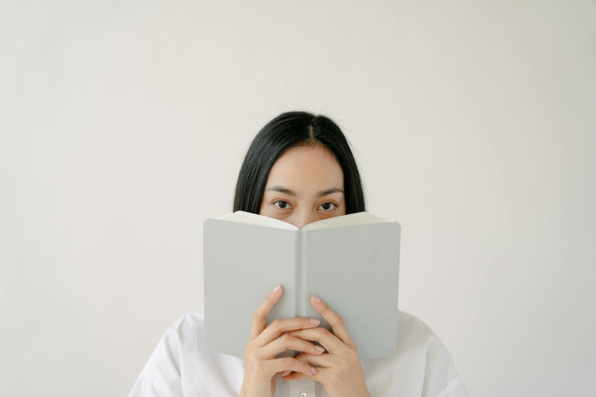 Jonge etnische vrouwelijke student met lang donker haar dat het gezicht bedekt met een notitieboekje terwijl ze zich voorbereidt op examens tegen een witte achtergrond