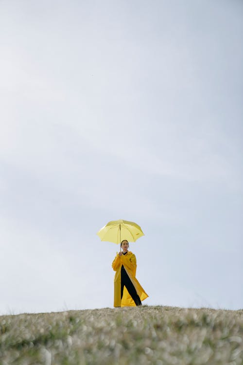 Low angle of unrecognizable female model in stylish yellow raincoat standing on grassy meadow with umbrella in hand against blue sky in hilly terrain