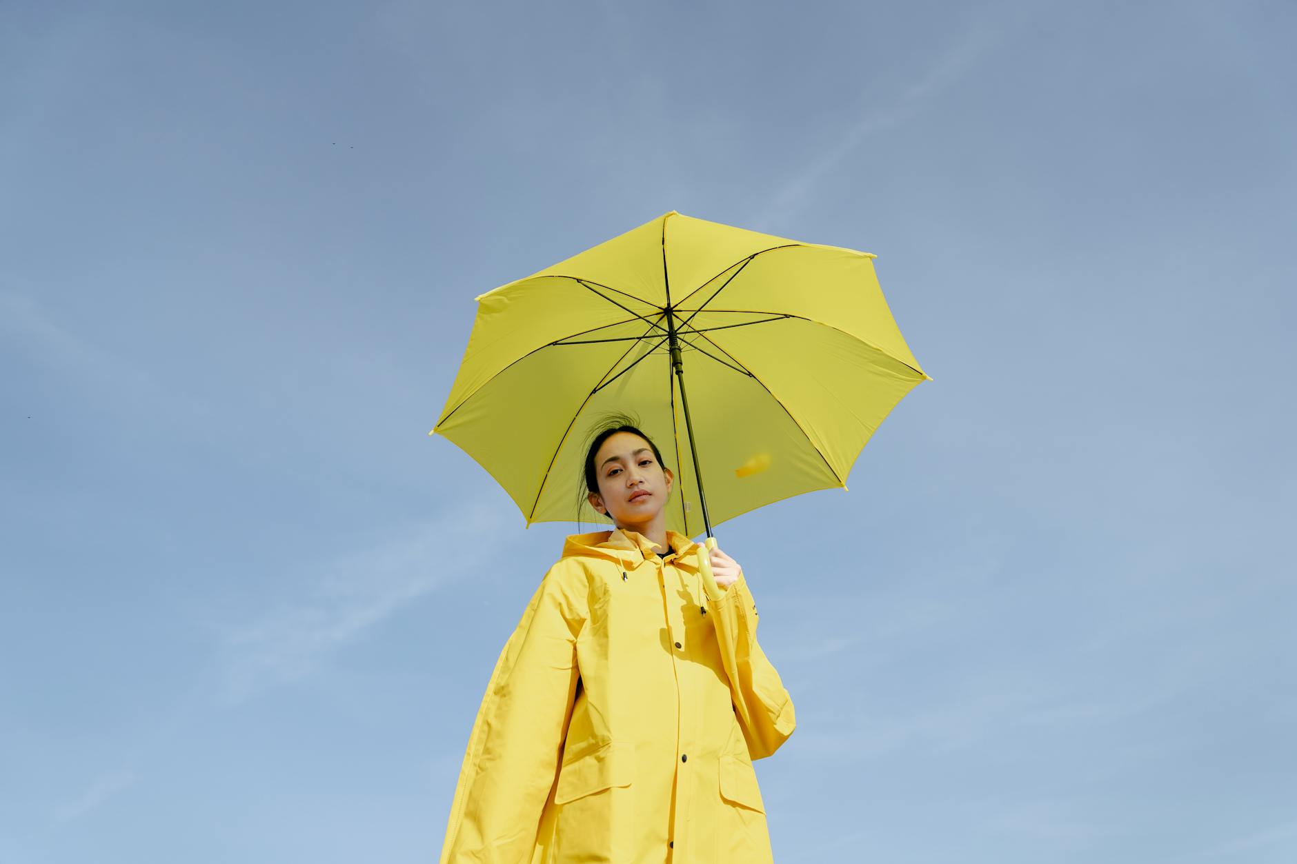 From below of trendy young Asian female with dark hair in bright raincoat and umbrella in hand looking at camera against blue sky on sunny day