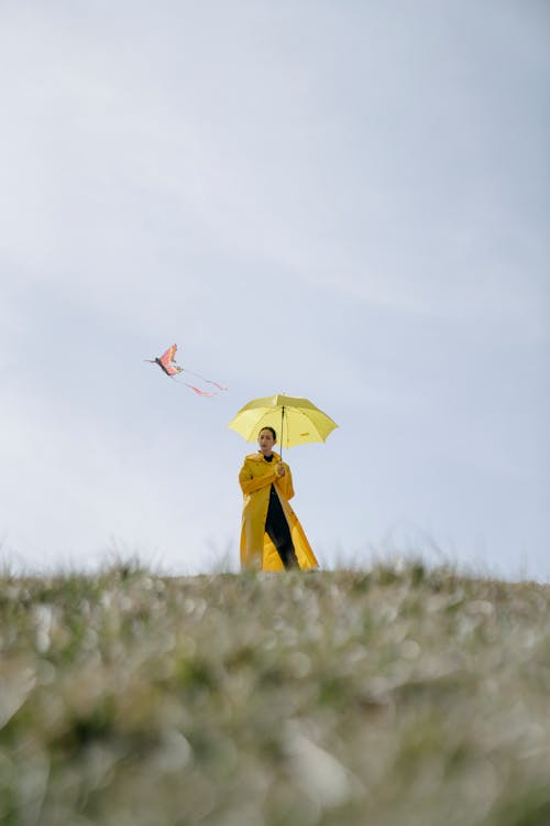 Stylish young woman with umbrella launching kite on grassy hill