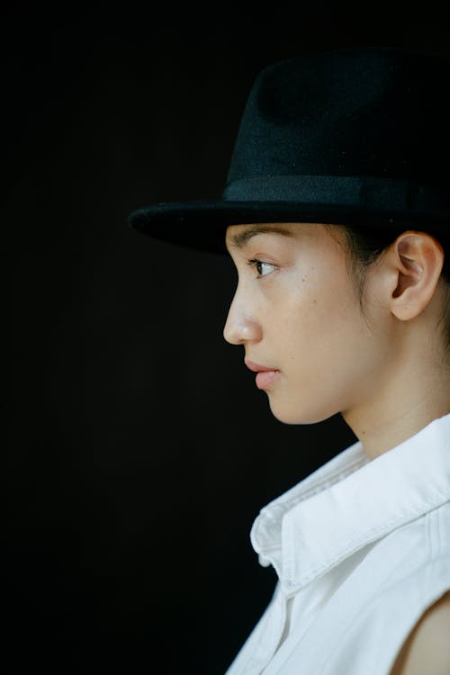 Side view of ethnic female wearing white sleeveless shirt and hat against black background