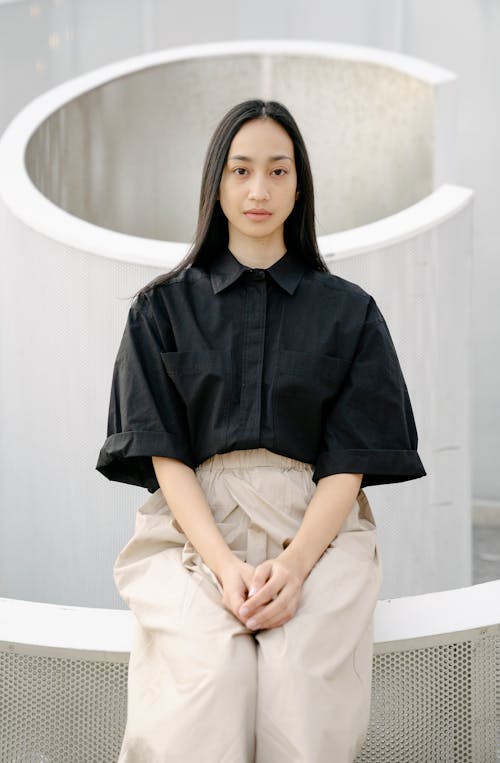 Emotionless ethnic female in shirt and pants sitting on fence and looking at camera