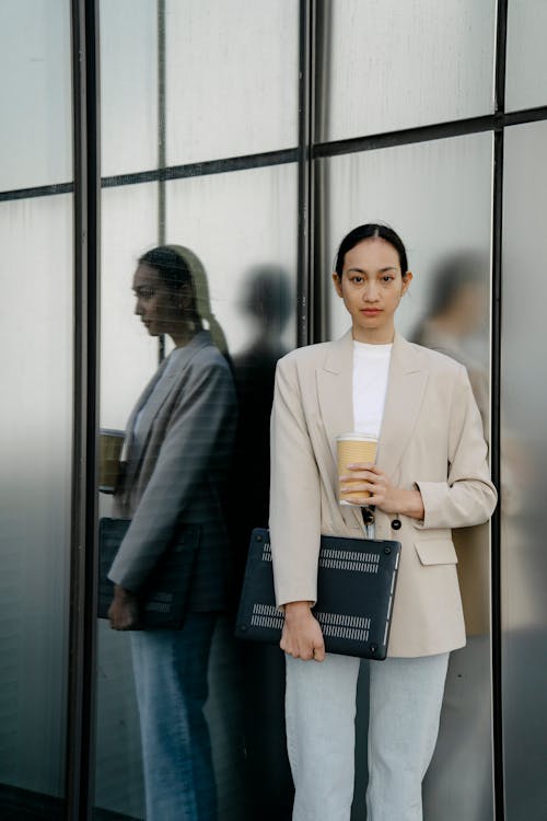 Free Confident ethnic female executive worker in formal wear standing with takeaway coffee and netbook near glass wall and looking at camera Stock Photo