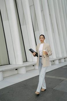 Asian woman with coffee and tablet walking outdoors, showcasing modern business style.