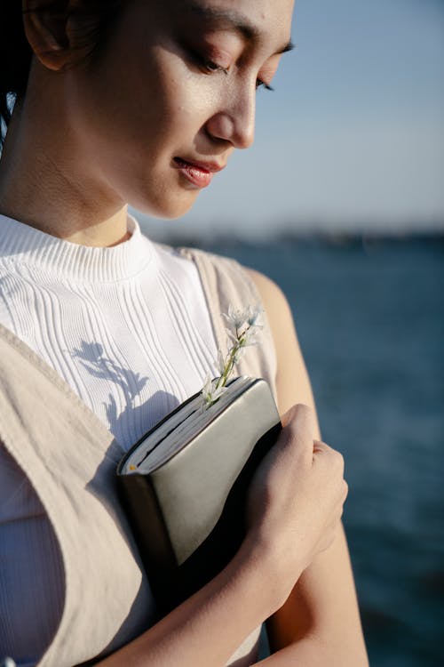 Dreamy ethnic woman embracing notebook on embankment