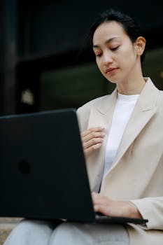 Concentrated Asian female entrepreneur in formal jacket typing on netbook while working on project remotely