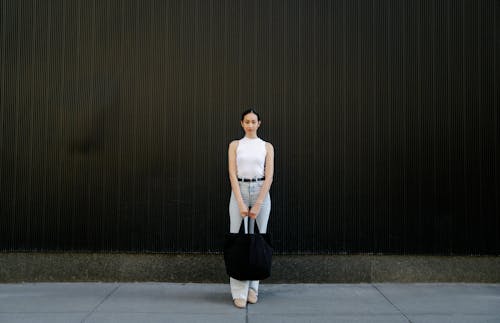 Full body of self assured young Asian lady with dark hair in trendy outfit standing on street near black wall with bag in hand and looking at camera