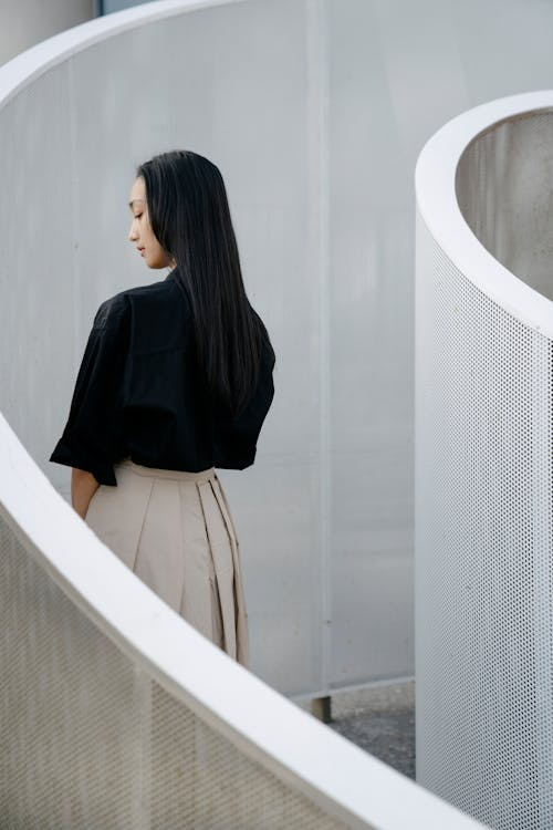 Free Back view of feminine young Asian lady with long dark hair in elegant outfit standing in curvy passage with metal walls and looking down pensively Stock Photo