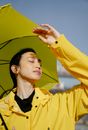 Dreamy young ethnic female with closed eyes in yellow raincoat enjoying sunlight under umbrella in town
