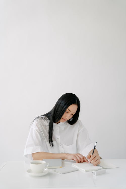 Attentive young ethnic female worker taking notes in journal at table with cup of coffee on light background