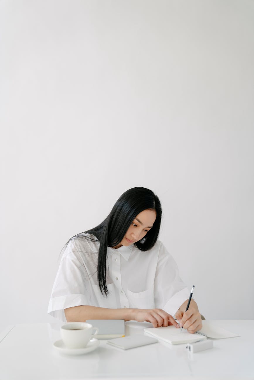 asian girl with white background sat at a table, writing on paper