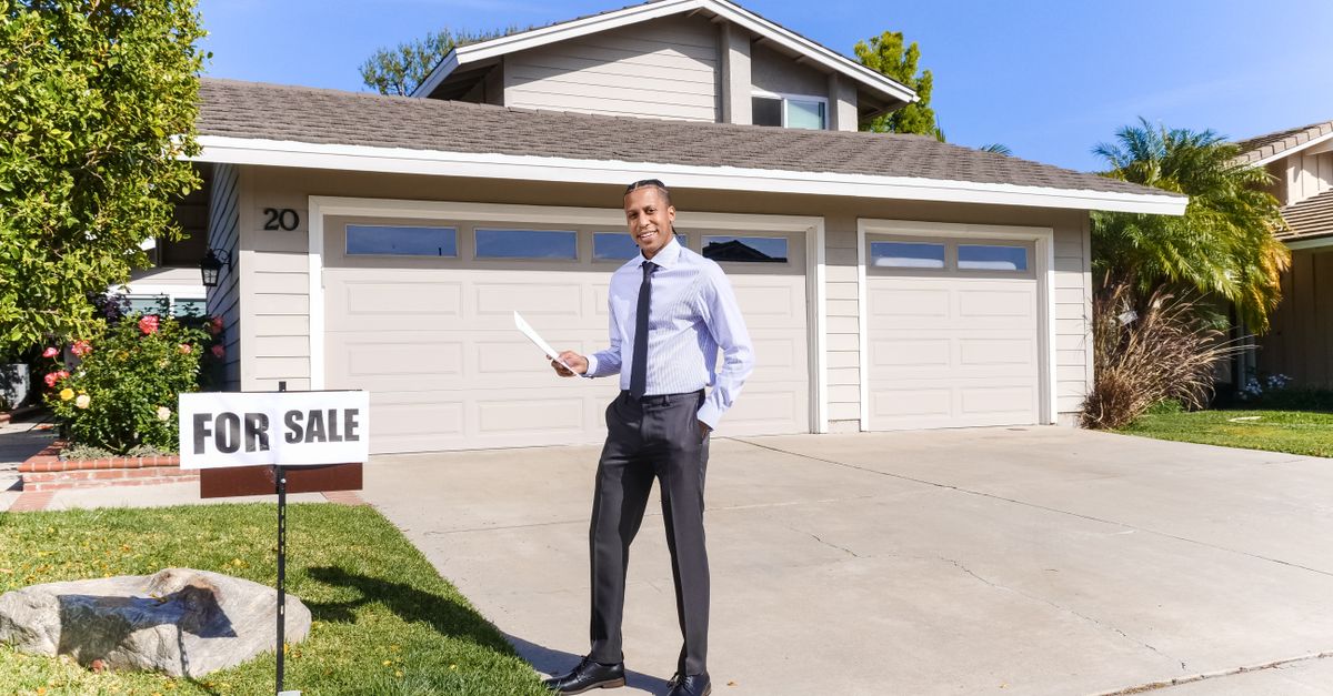 Real estate agent standing outside a house with a for sale sign in the driveway.
