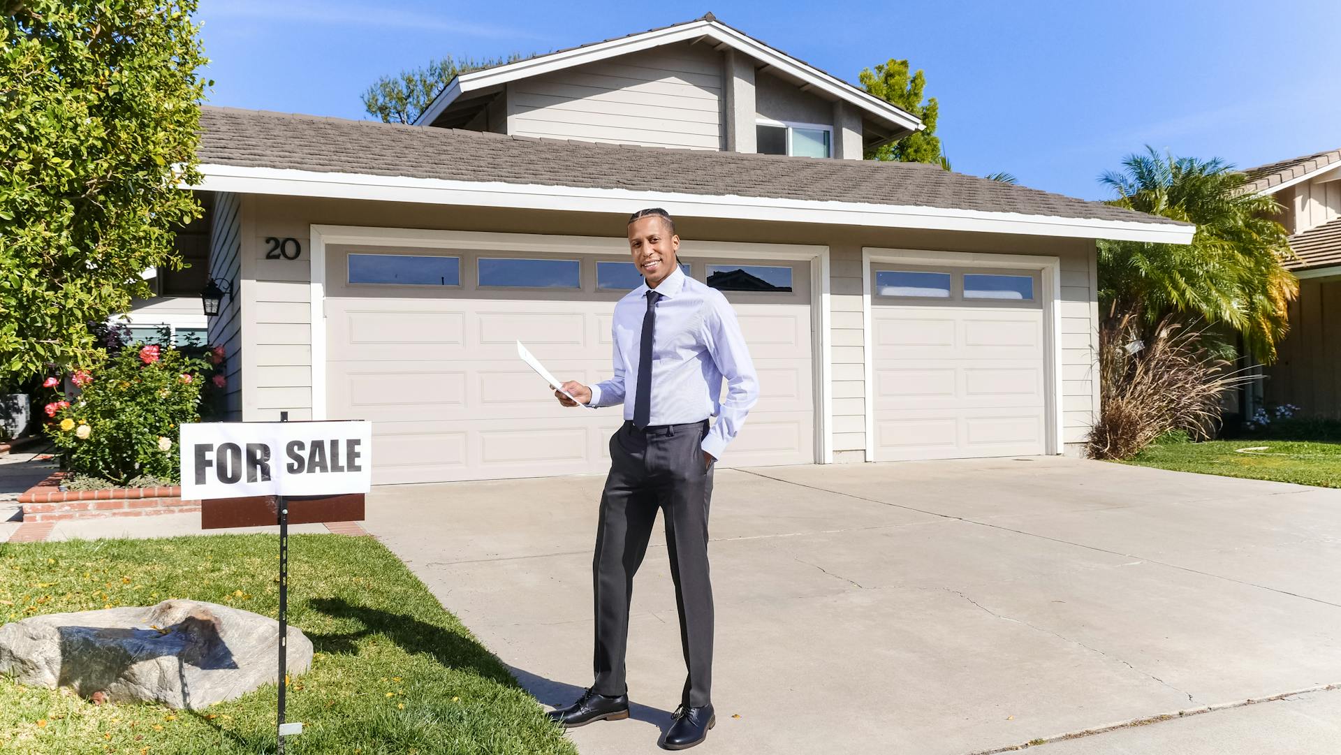 Real estate agent standing outside a house with a for sale sign in the driveway.