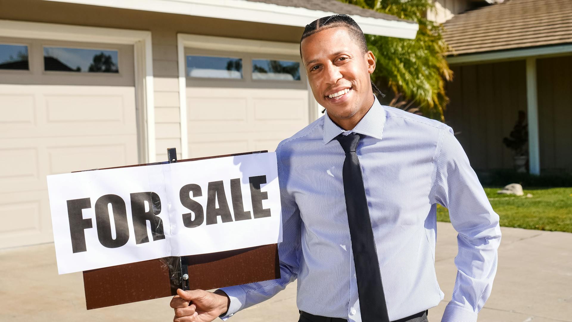 Real estate agent standing with a 'For Sale' sign in front of a house on a sunny day.