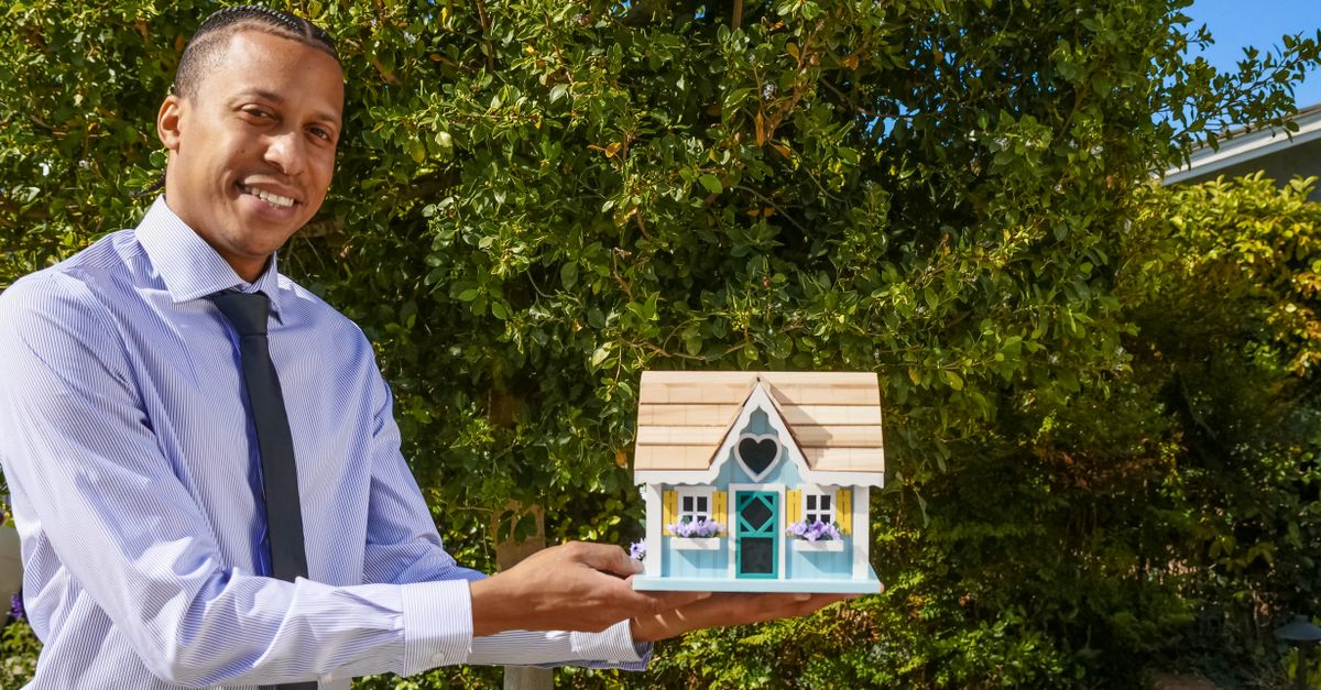 Smiling real estate agent presenting a miniature house model outdoors against a background of greenery.
