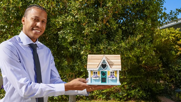 Smiling real estate agent presenting a miniature house model outdoors against a background of greenery.