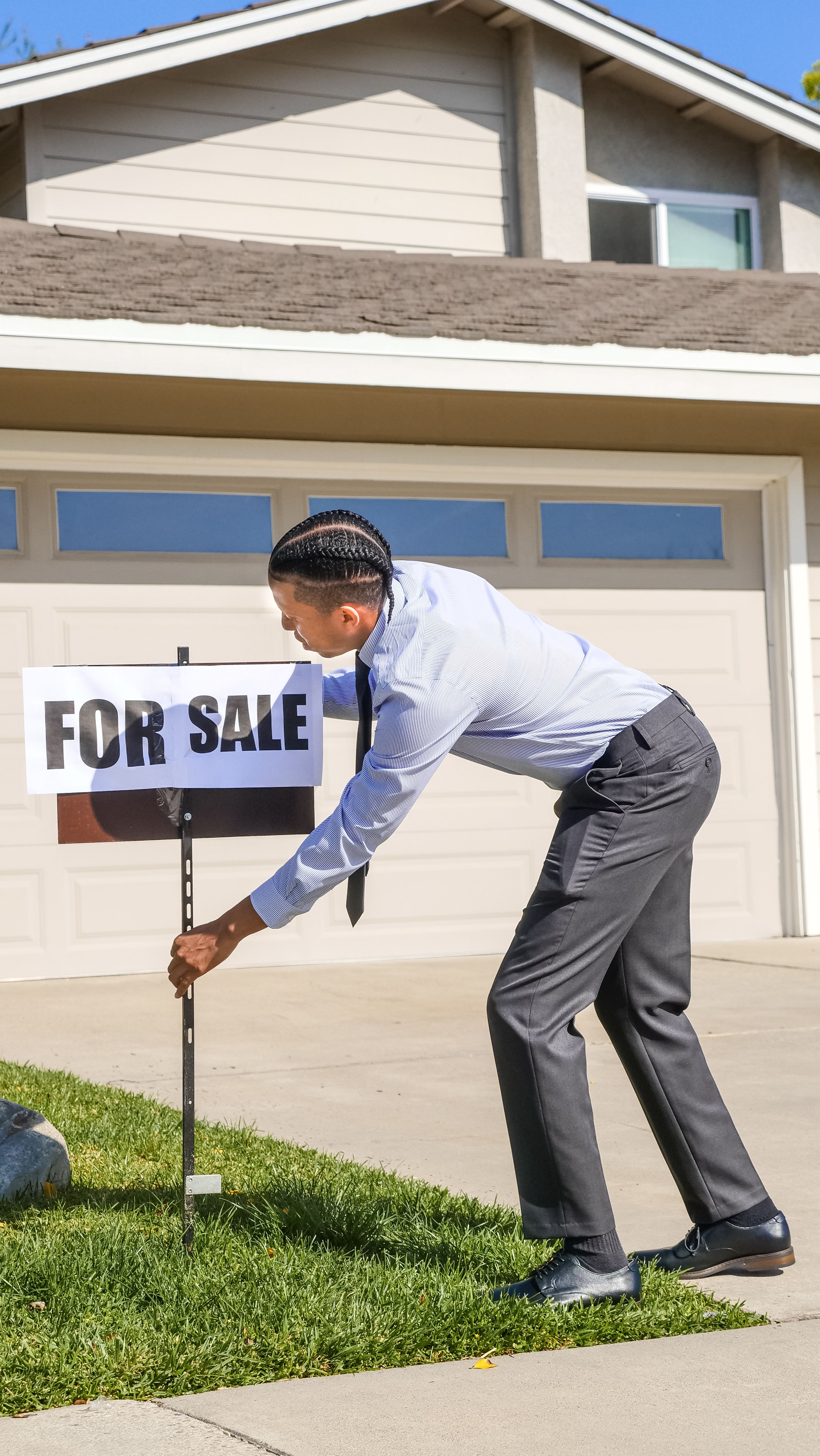 Alt Text: A male real estate professional wearing a dark shirt placing a white and red "For Sale" yard sign into the green grass of a suburban lawn.