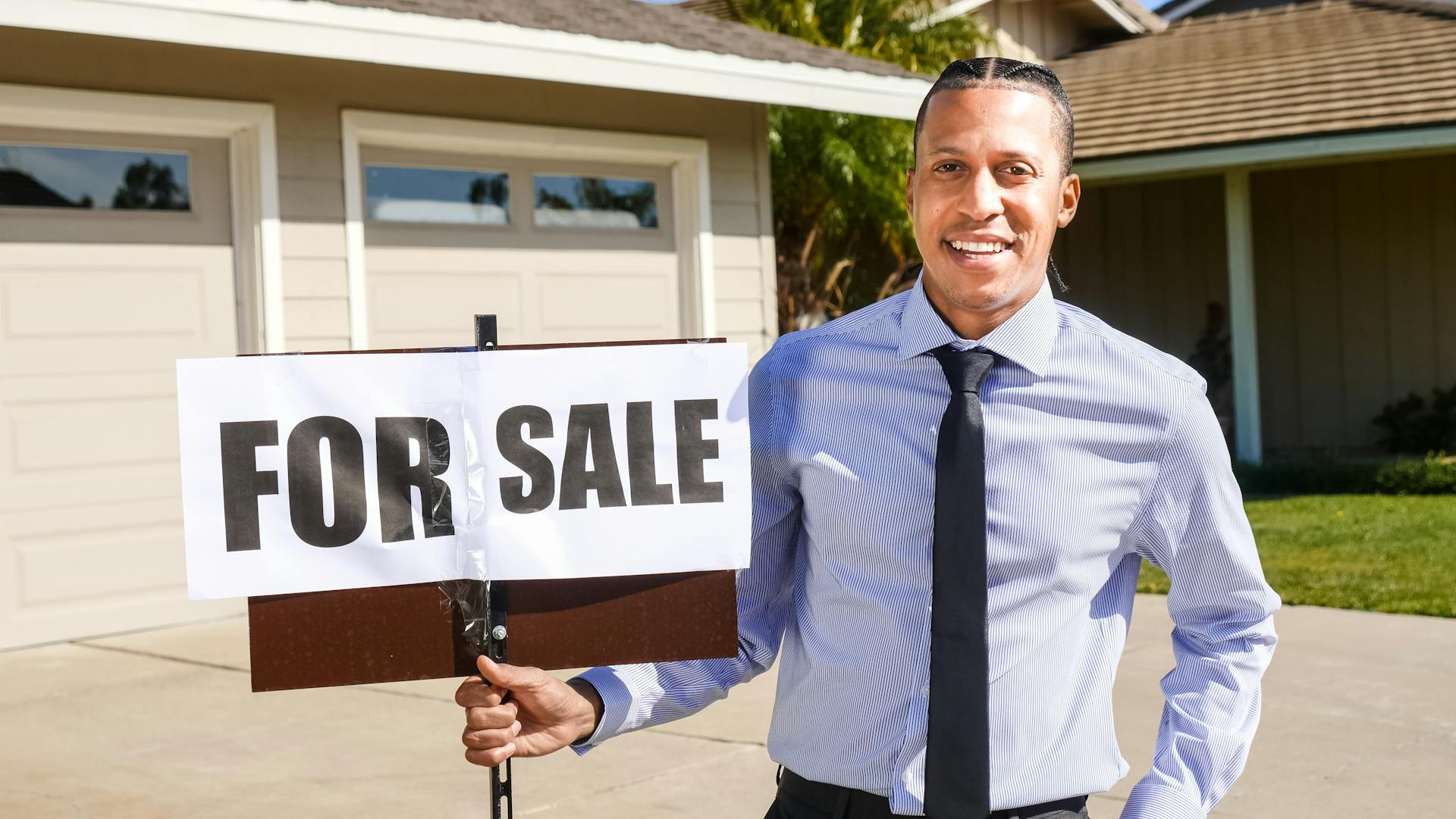 Smiling real estate agent with a for sale sign in front of a house.