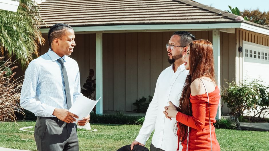 A real estate agent discusses property details with a couple outside a suburban home.