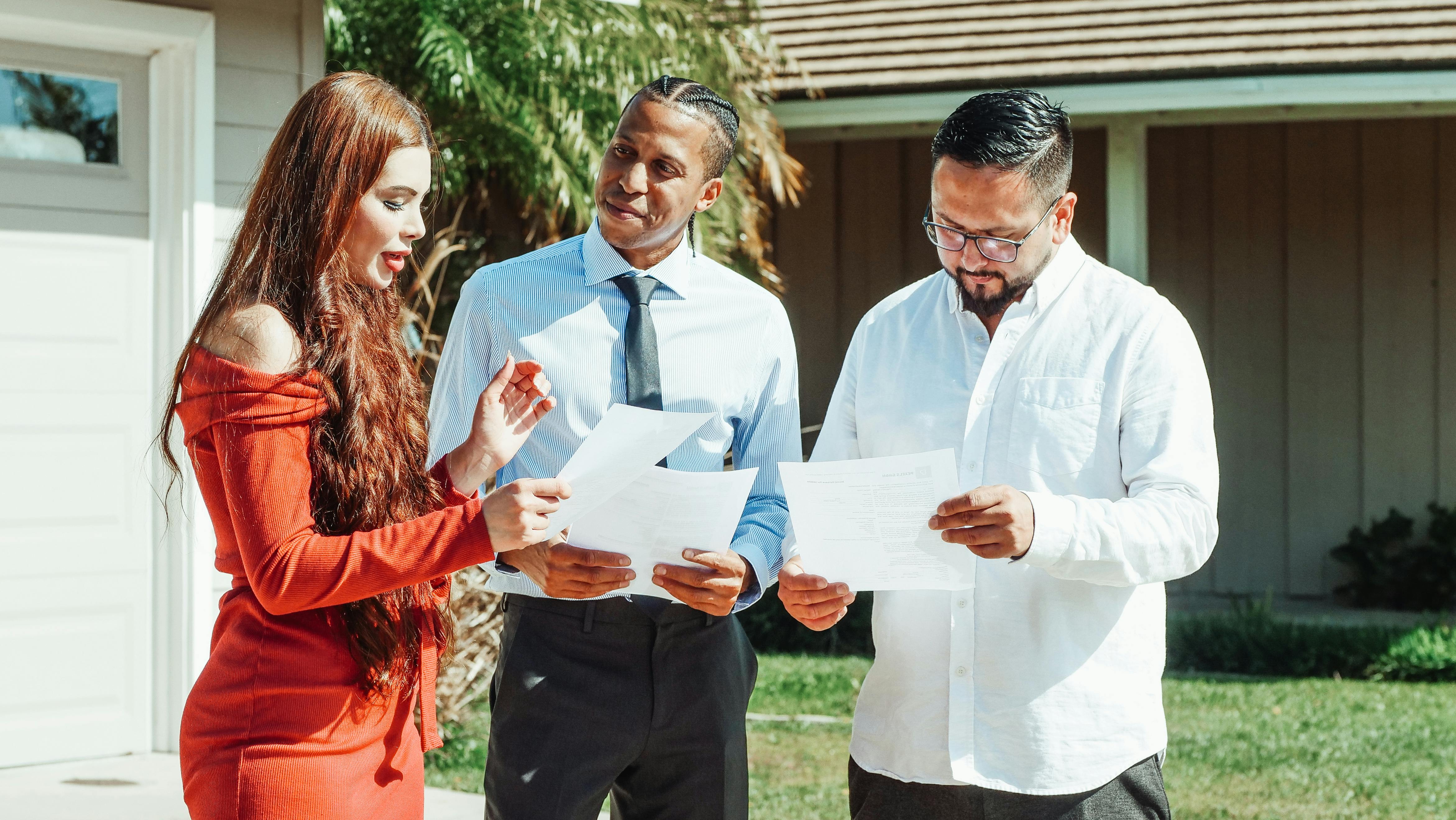 A diverse group evaluates real estate documents outside a residential property in daylight.