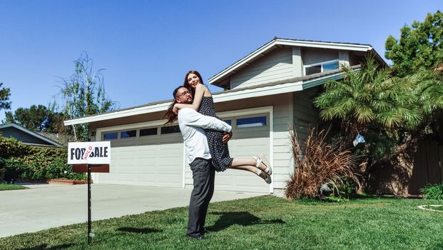 A joyful couple celebrating their new home purchase in front of a house with a sold sign.