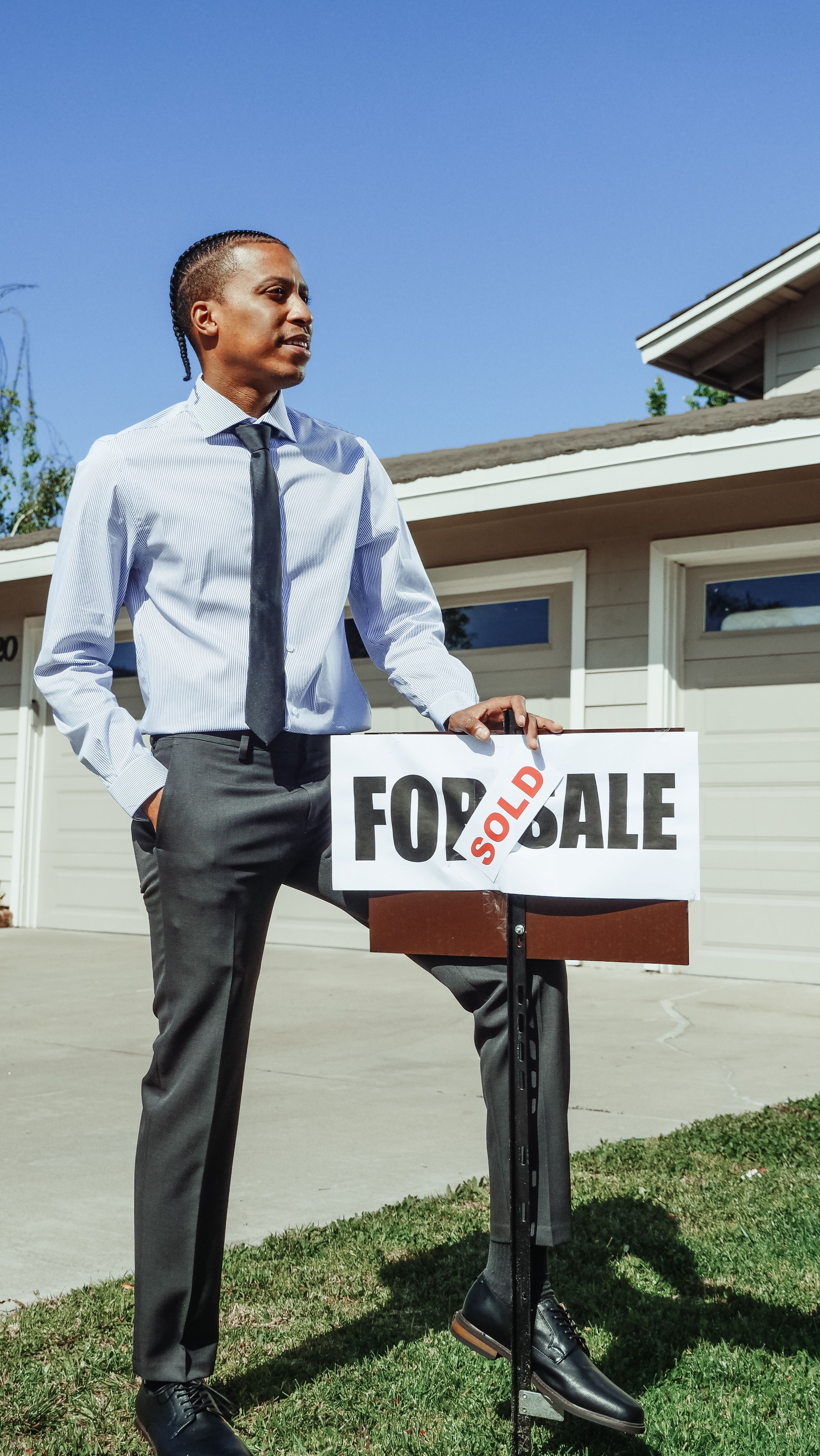 A smiling man dressed in a blue dress shirt standing on a suburban property holding a bright red "SOLD" sign.