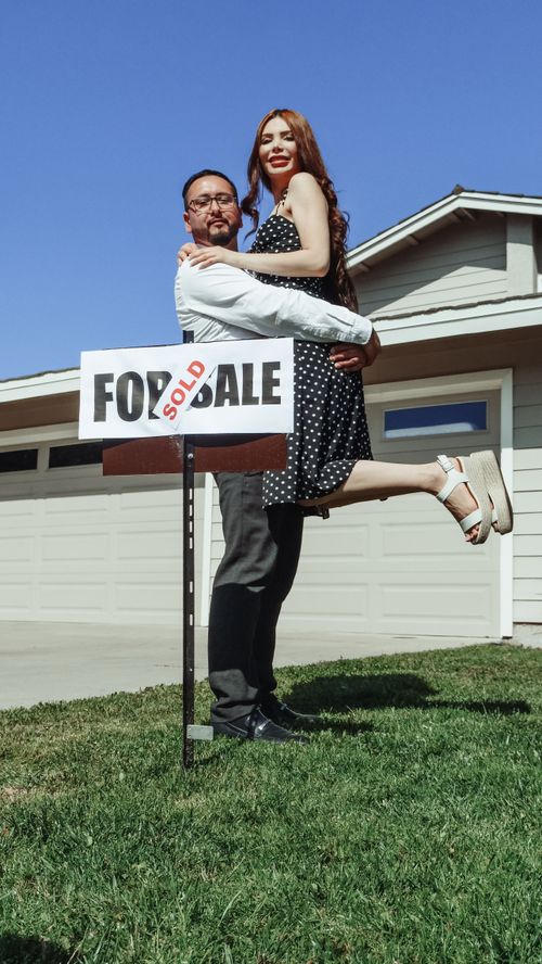 Free A joyful couple celebrates buying a new home as they pose with a sold sign in front of a house. Stock Photo